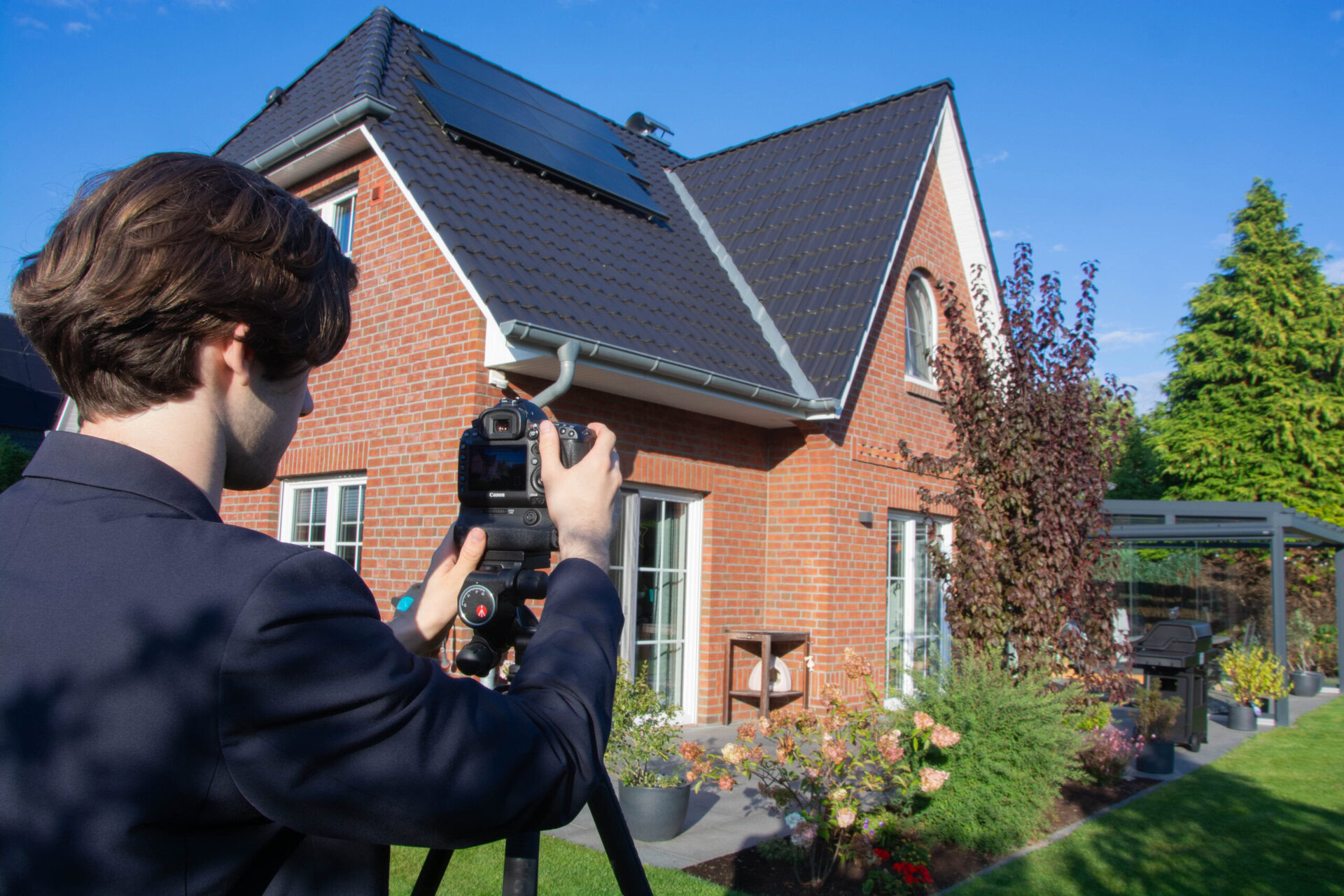Ein Immobilienmakler fotografiert ein modernes Einfamilienhaus mit Garten in sonnigem Wetter.